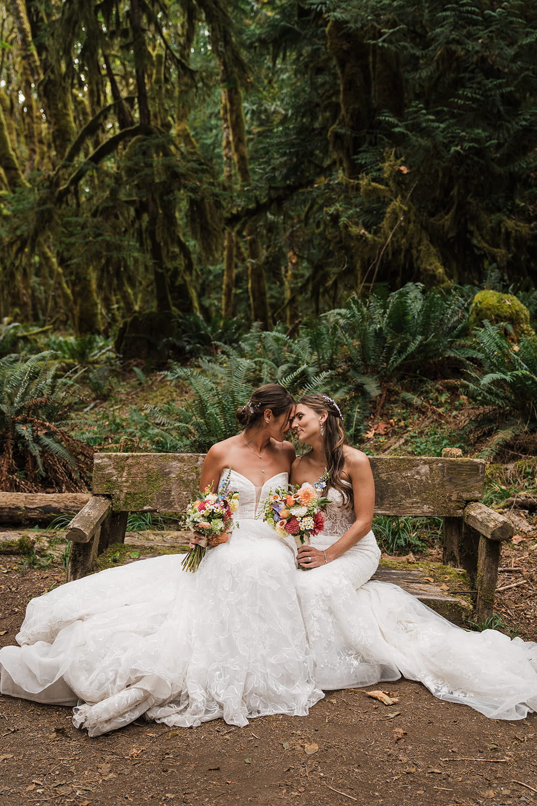 Brides sit on a mossy bench in the forest in their wedding dresses.