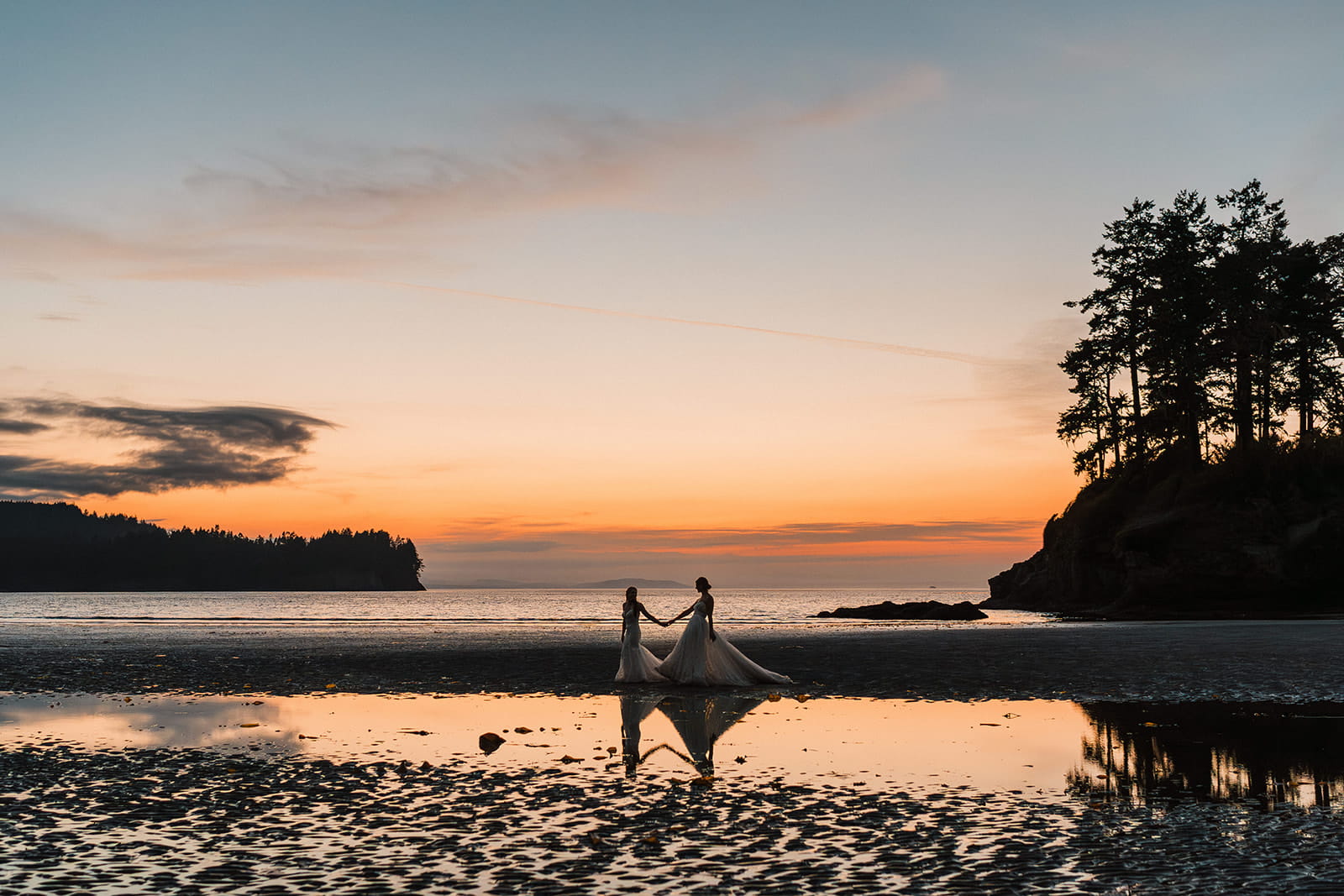 Brides hold hands and walk across the beach after sunset. 